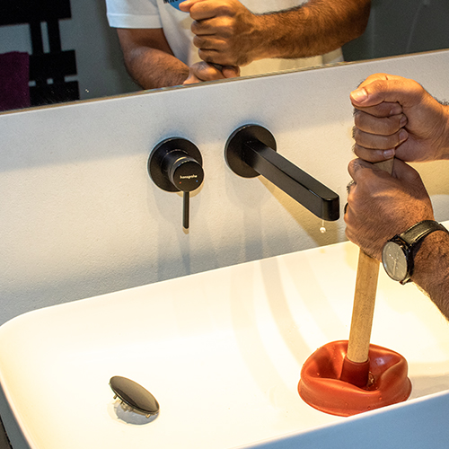 Ein Mitarbeiter von Sauber In Form benutzt einen Pümpel, um ein Waschbecken zu reinigen und eine Verstopfung zu beseitigen.