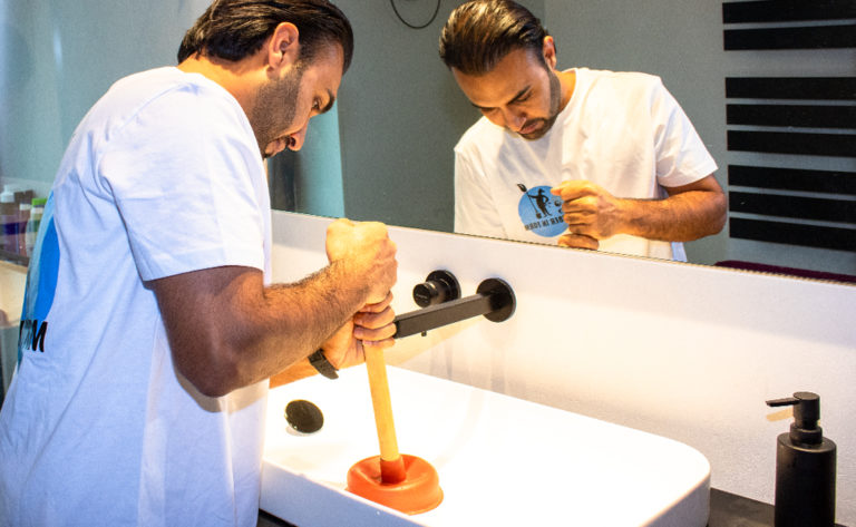 Ein Mitarbeiter von Sauber In Form beseitigt im Badezimmer eine Verstopfung im Waschbecken mit einem Pümpel.