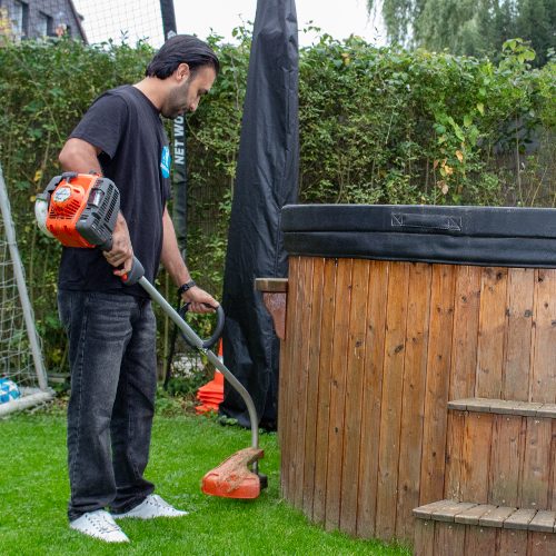 Ein Mitarbeiter von Sauber In Form trimmt sorgfältig das Gras rund um einen Holzwirlpool im Garten.