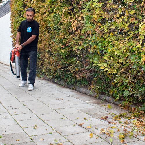 Ein Mitarbeiter von Sauber In Form entfernt Herbstlaub mit einem Laubbläser von einem gepflasterten Weg neben einer Hecke.