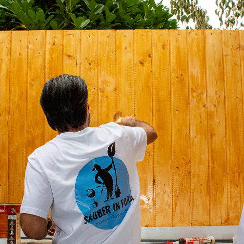 Ein Hausmeister von Sauber In Form streicht eine hölzerne Gartenwand mit frischer Farbe.